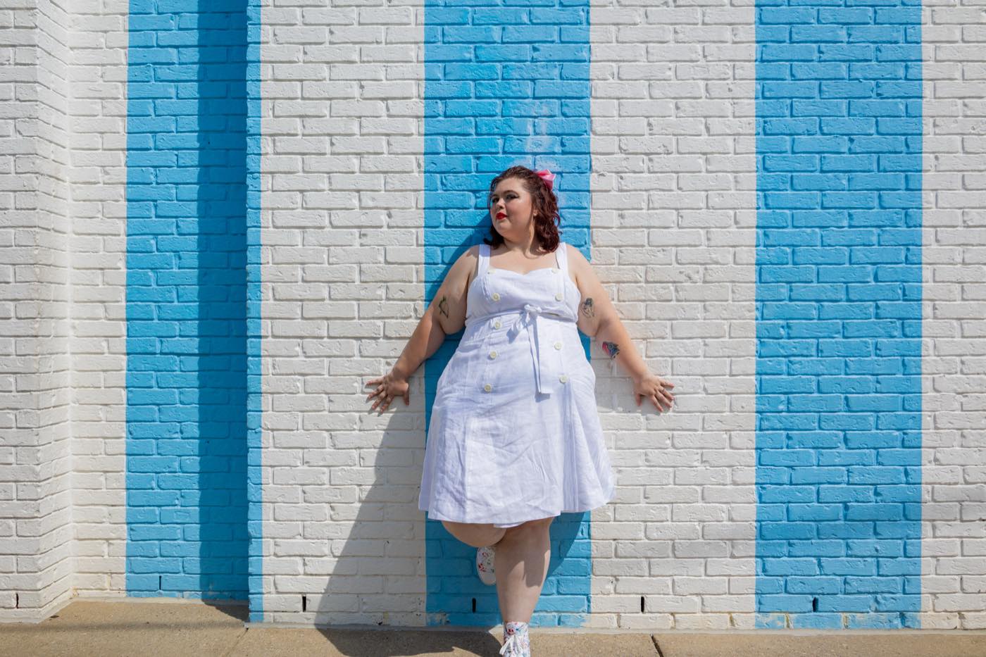 Katherine posing in a white dress against a blue and white striped brick wall