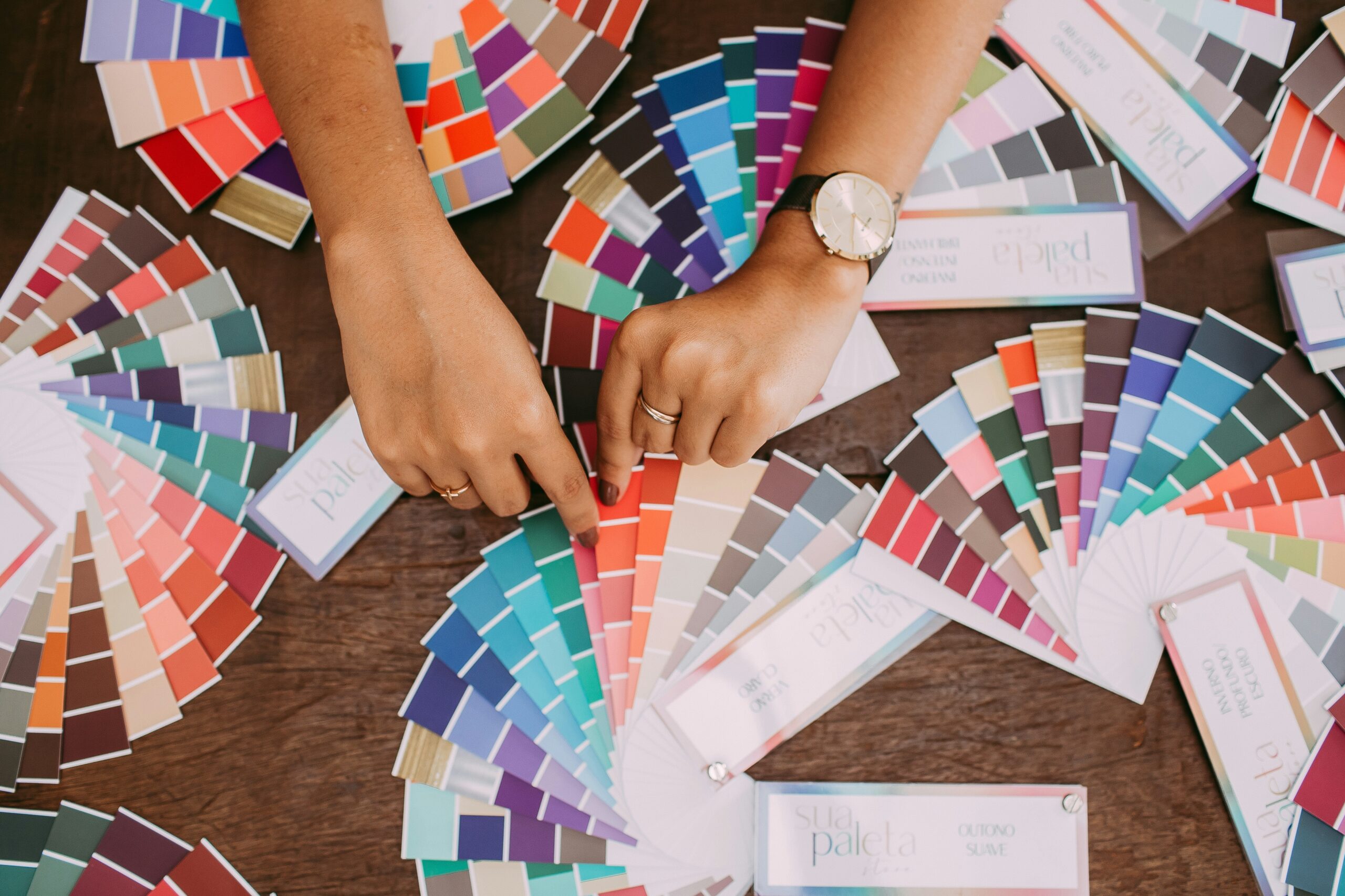 Katherine Magpie Design — Two hands point at colored paint swatches spread out on a wooden table, thoughtfully considering palettes for web design for romance authors. One hand wears a watch and a ring, while various palettes display an array of vibrant shades in a fan arrangement. — Web design tips for romance authors